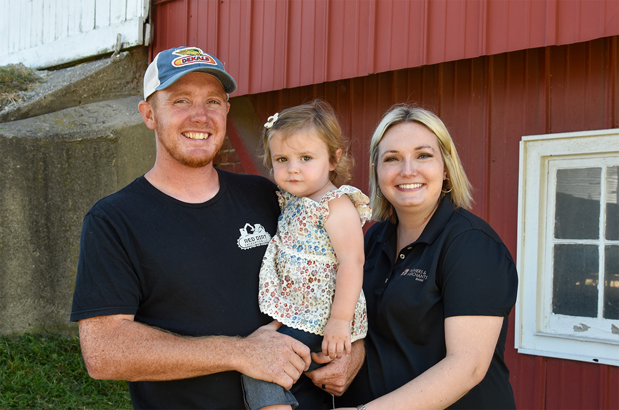 Family smiling at the camera in front of a red building.